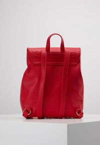 Mochila roja hecha de un material sintético suave, con una forma estructurada, correas ajustables y un diseño minimalista. Cuenta con una tapa superior.