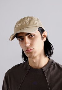 Tonal beige baseball cap with embroidered text "sm*Je." Worn with a dark-colored sweatshirt, featuring a round neckline and minimal design.