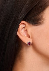 Silberohrringe mit einem herzförmigen roten Edelstein, umgeben von kleinen klaren Steinen, vor glattem braunem Haar und Haut.