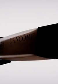 Lancôme foundationflaske med matt svart hette, slank og forlengt form, og preget merkenavn på den beige kroppen.