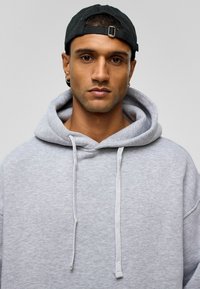 Grijze hoodie met een grote voorzak, trekkoorden en een brede kraag. Draag onder een zwarte baseballcap. Glad van structuur. Neutrale achtergrond.