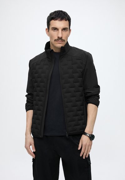 Homme aux cheveux courts et foncés avec une moustache, portant une veste matelassée noire sur une chemise noire et un pantalon noir, sur un fond uni clair.