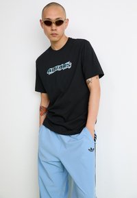 T-shirt de algodão preto com logotipo da Adidas em teal e cinza na frente, combinado com calças de poliéster azuis claras com emblema preto da Adidas.