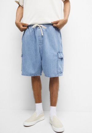 Personne portant un short cargo en denim bleu clair avec taille à cordon, des chaussettes blanches, des chaussures beige à enfiler et une chemise blanche à manches courtes.