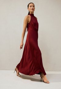 Maxikjole i en dyb bordeauxfarve med høj hals, ærmeløs design og en let, flydende tekstur. Kompletteret med beige højhælede sandaler.