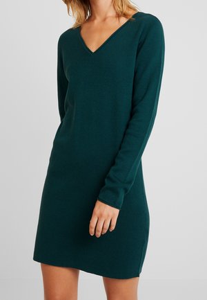 Femme portant une robe en maille vert foncé à manches longues avec un col en V, debout devant un fond clair uni.