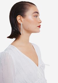 Lange sølvøreringe med kaskader af krystalstrenge. Båret af en person med kort, glat tilbagetrukket hår, iført en hvid kjole med sølvdetaljer.