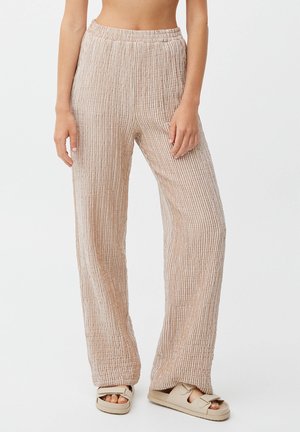 Femme portant un pantalon beige côtelé à jambes larges et des sandales beiges à bouts ouverts, debout devant un fond uni.