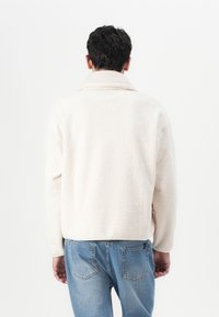 Cream textured fleece jacket with a wide collar and ribbed accents, paired with light blue denim jeans. Visible seams and relaxed fit.