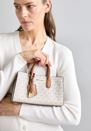 Kobieta w białym swetrze trzyma beżową i brązową torebkę Michael Kors z monogramem i złotym zamkiem na jednolitym tle.