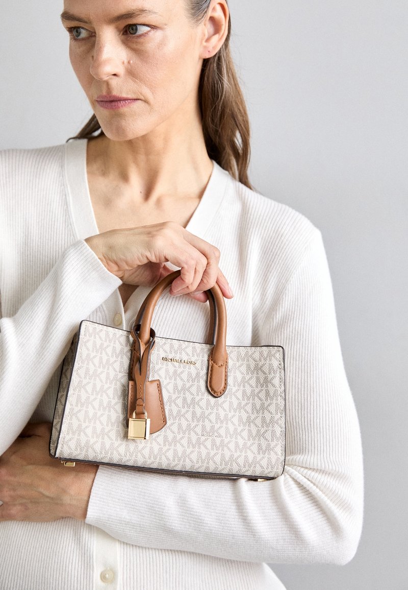 Femme en pull blanc tenant un sac à main Michael Kors beige et marron avec motif monogramme et détail de verrou doré sur un fond uni.