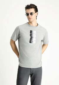 Camiseta de algodón gris con un diseño de texto en negro y blanco en negrita. Modelo vistiendo pantalones grises y gafas de sol redondas. Textura suave, ajuste casual.