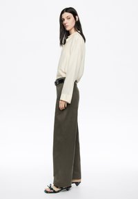 Maglietta lunga crema con colletto, abbinata a pantaloni larghi color oliva. Una cintura nera e sandali con tacco decorati da un motivo floreale completano l'outfit.