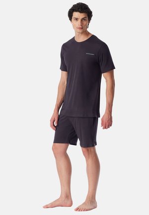 Młody mężczyzna stoi boso, ubrany w ciemny t-shirt z krótkim rękawem i dopasowane szorty z małym logo na piersi.