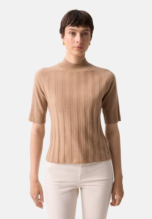 Kvinde med kort mørkt hår iført beige ribstrikket kortærmet sweater og hvide bukser, stående mod en ensfarvet baggrund.
