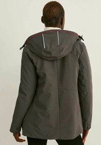 Voděodolný šedý kabát s kapucí, s červenými akcenty a nastavitelnými prvky. Hladká textura s přiléhavým střihem a strukturovaným designem.