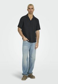 Schwarz kurzärmliges Hemd mit Knopfverschluss und strukturiertem Muster, kombiniert mit hellblauen Jeans und braunen Slippers. Heller Hintergrund.