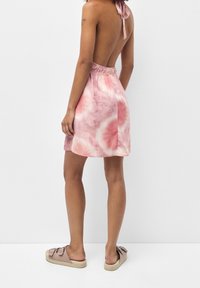 Mujer con un vestido mini sin espalda en tonos rosa y blanco con un diseño de tie-dye, llevando sandalias deslizables, delante de un fondo blanco plano.