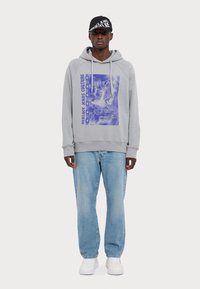 Felpa grigia con cappuccio e stampa grafica viola, abbinata a jeans azzurri chiari e sneakers bianche; il modello indossa un cappellino nero con del testo.