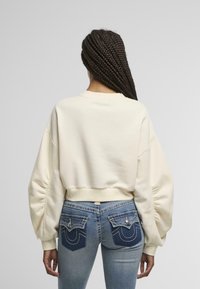 Sudadera cortada de color crema con mangas voluminosas, combinada con jeans azules de tiro medio que presentan bolsillos traseros decorativos y acentos de costura.