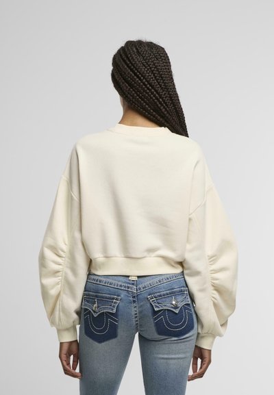 Sweatshirt crème coupé avec des manches volumineuses, associé à un jean bleu taille mi-haute avec des poches arrière décoratives et des accents de couture.