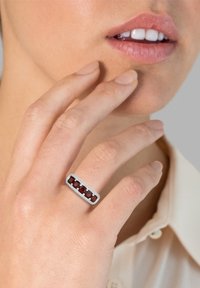 Rechteckiger Silberring mit fünf tiefroten Edelsteinen, in einem strukturierten Band mit feinen Details gefasst, der an einer Hand getragen wird, die eine Wange berührt.