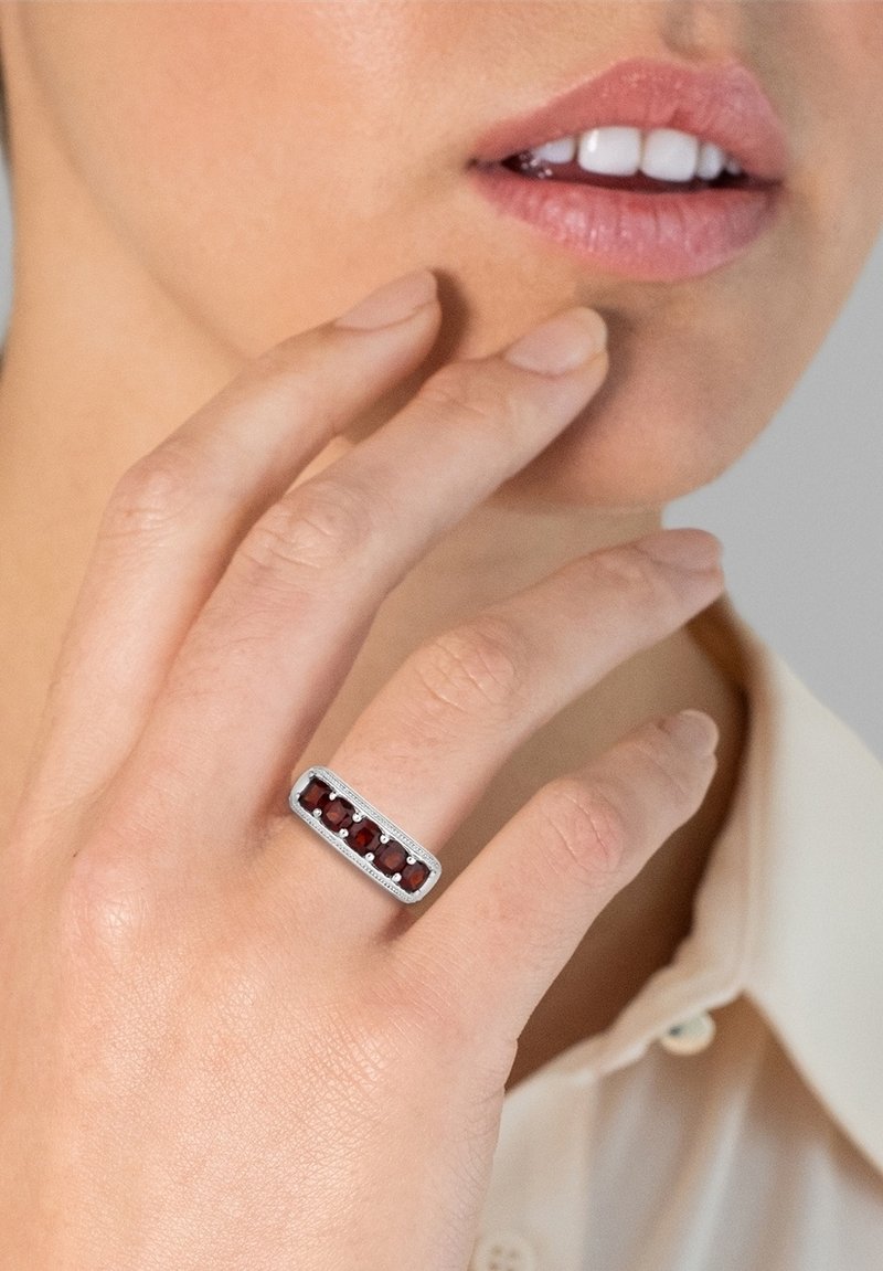 Rechteckiger Silberring mit fünf tiefroten Edelsteinen, in einem strukturierten Band mit feinen Details gefasst, der an einer Hand getragen wird, die eine Wange berührt.