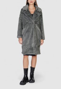 Cappotto in pelliccia sintetica grigia con ampio colletto, chiusura a doppio bottone e maniche lunghe. Indossato sopra un vestito nero e abbinato a stivali neri alla caviglia.
