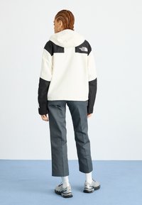 White and black fleece jacket with a hood, featuring a logo on the upper back, paired with gray trousers and sleek athletic shoes.