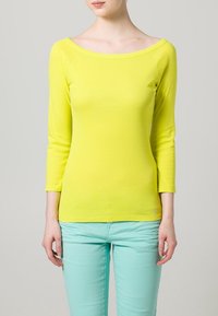 Žlutý top s 3/4 rukávy a lodním výstřihem, s hladkým povrchem. Spárovaný s mentolově zelenými slim kalhotami.