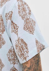 Chemise à manches courtes en tissu bleu clair ornée d'un motif floral marron. La texture est côtelée, avec une coupe décontractée et une finition douce.