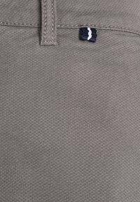 Gros plan sur un tissu gris texturé avec des coutures et une petite étiquette tissée bleu marine et blanche près d'une passants de ceinture.