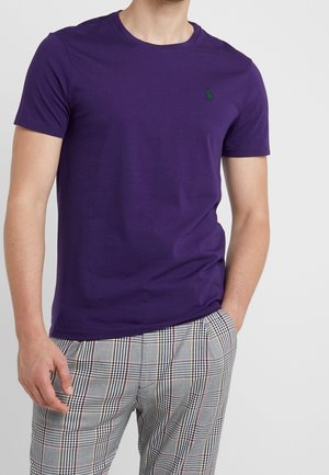 Mann, der ein schlichtes lila T-Shirt mit kurzen Ärmeln und graue karierte Hose trägt, eine Hand in der Tasche, vor einem einfarbigen Hintergrund.