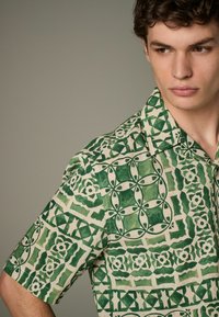 Chemise à manches courtes verte et crème avec un motif géométrique. Réalisée en tissu léger avec des motifs floraux et abstraits.