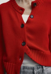 Röd stickad kofta med rund halsringning, svarta knappar och kort passform. Har texturerat tyg och en detaljerad slits på sidan.