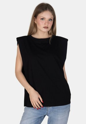 Giovane donna con capelli lunghi indossa un top nero senza maniche e jeans azzurri, posando con una mano sul fianco contro uno sfondo bianco.