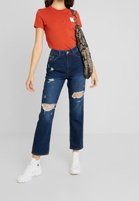 Röd åtsittande t-shirt, högmidjade mörkblå slitna jeans med rivna detaljer. Vita sneakers och en svart texturerad axelväska.