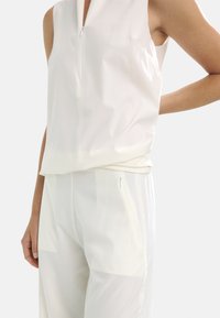 Biały, bezrękawnik z zamkiem błyskawicznym z przodu, wykonany z gładkiego, lekkiego materiału. Dolny brzeg jest nieco luźny, doskonale współgrając z dopasowanymi, przezroczystymi spodniami.