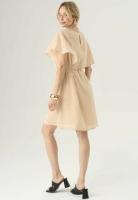 Abito beige pallido con maniche a sbuffo e vita legata, con dettaglio di apertura a chiave sulla schiena. Abbinato a sandali neri con tacco alto.