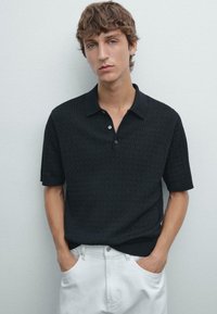 Polo shirt nera a maniche corte con motivo testurizzato e colletto, abbinata a pantaloni bianchi. Quattro bottoni visibili sul collo.