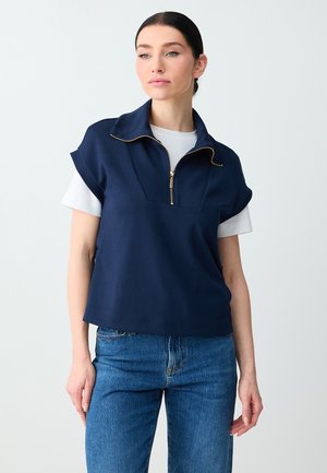 Vrouw draagt een marineblauw shirt met korte mouwen en rits, gedragen over een wit T-shirt en blauwe jeans, tegen een effen lichte achtergrond.