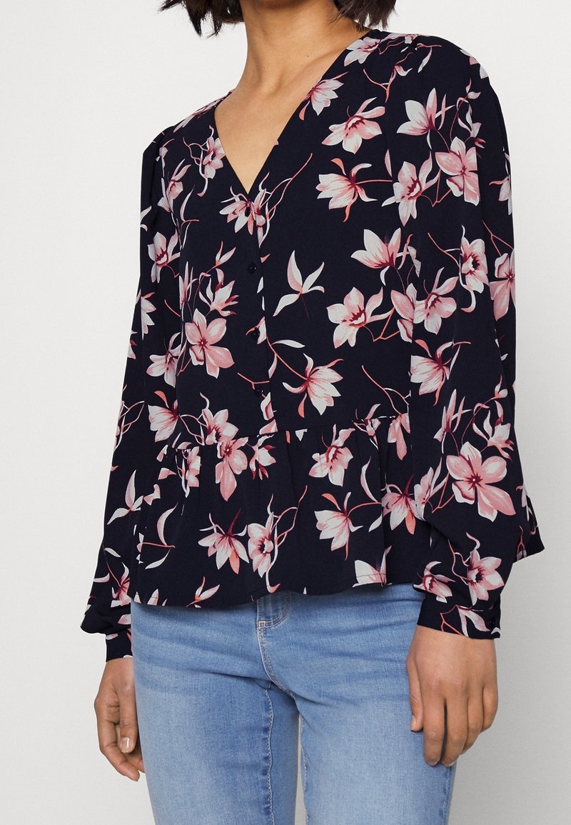 Femme portant un chemisier bleu marine avec un imprimé floral rose et blanc et des manches longues, associé à un jean bleu clair.