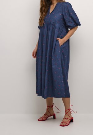 Denimkleid mit kurzen Puffärmeln, V-Ausschnitt und roten Herzstickereien. Fließendes Design mit seitlichen Taschen. Kombiniert mit roten Riemchensandalen.
