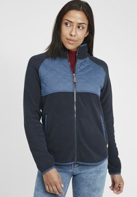 Veste en polaire marine avec un empiècement matelassé bleu clair, fermeture éclair intégrale, poches zippées et bordures bleues le long des bords. Caractérisée par un col montant.