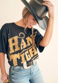 Mustapohjainen ylisuuri t-paita, jossa on suuret kultaiset tekstit "HANG TIGHT", yhdistettynä vaaleansinisiin korkeavyötäröisiin farkkuihin ja leveälieriseen mustaan hattu.