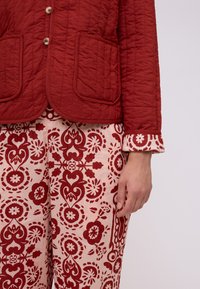 Giacca trapuntata rossa con due tasche frontali, abbinata a pantaloni rossi e crema a fantasia con motivi floreali e orli arrotolati.