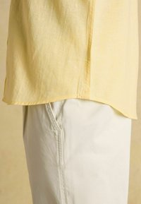 Jasnożółty len, dół koszuli na kremowych spodniach przy kieszeni, zbliżenie ukazujące fakturę tkaniny i szczegóły szwów.