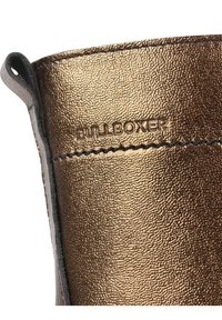 Gros plan sur la tige de botte en cuir bronze métallique avec surpiqûres noires et nom de marque embossé "PULL&BOXER."