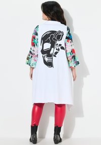 Giacca bianca lunga con una grafica di teschio e rosa nera sulla schiena, maniche a fantasia multicolore, leggings rossi e stivali neri con borchie.