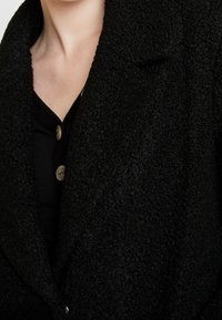 Manteau texturé noir avec col châle, présentant une apparence douce et duveteuse ainsi qu'un léger éclat, fermé par des boutons de couleur claire.